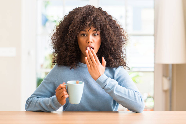 Африканская американка, держащая чашку кофе у себя дома, прикрывает рот рукой, потрясенной стыдом за ошибку, выражением страха, испуганной в тишине, секретной концепцией
 - Фото, изображение
