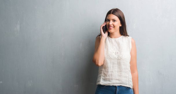 Young brunette woman over grunge grey wall talking on the phone with a happy face standing and smiling with a confident smile showing teeth - Zdjęcie, obraz