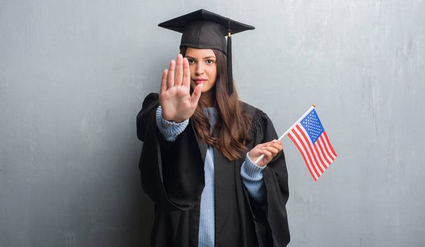グランジの若いブルネットの女性灰色の壁の防衛のジェスチャー、深刻な自信を持って式に一時停止の標識を行うオープンの手でアメリカの旗を保持している大学院の制服を着て - 写真・画像
