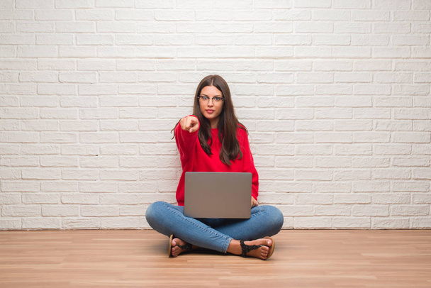 Nuori brunette nainen istuu lattialla yli valkoinen tiili seinään kannettavan tietokoneen sormella kameraan ja sinulle, käsi merkki, positiivinen ja luottavainen ele edestä
 - Valokuva, kuva