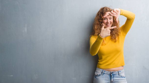 Jonge roodharige vrouw over grijs grunge muur glimlachend maken frame met handen en vingers met blij gezicht. Creativiteit en fotografie concept. - Foto, afbeelding