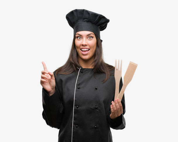 Młodzi Hiszpanie gotować kobieta ubrana chef uniform zaskoczony z pomysłu lub pytanie palcem wskazującym z szczęśliwy twarz, numer jeden - Zdjęcie, obraz
