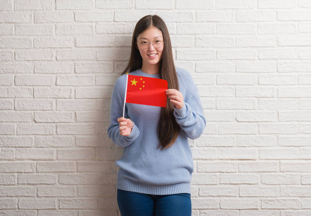 Fiatal kínai nő gazdaság Kína zászlaja, boldog arccal állt, és mosolyogva, magabiztos mosollyal mutatja a fogak tégla falon - Fotó, kép