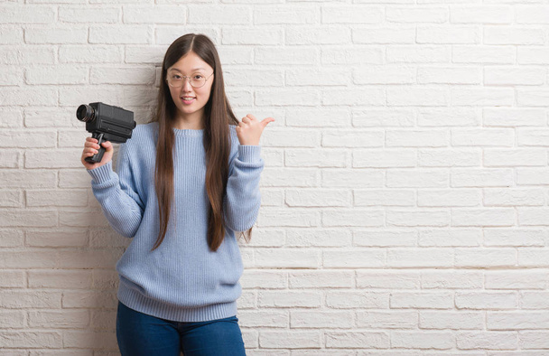 Nuori kiinalainen nainen kuvaamisen pitäen videokamera osoittaa ja osoittaa peukalolla sivulle iloinen kasvot hymyillen
 - Valokuva, kuva