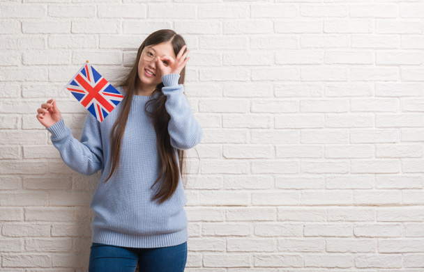 junge Chinesin über Ziegelmauer hält Flagge von England mit glücklichem Gesicht lächelnd tut ok Zeichen mit Hand auf Auge durch Finger schauen - Foto, Bild