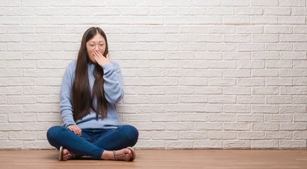 junge Chinesin sitzt auf dem Boden über Ziegelwand gelangweilt gähnen müde Mund mit der Hand bedeckt. Ruhelos und schläfrig. - Foto, Bild