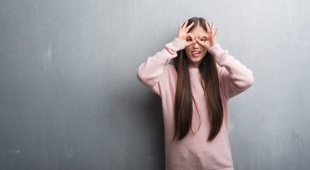 Giovane donna cinese sopra il muro grigio facendo gesto ok come binocolo sporgere la lingua, occhi che guardano attraverso le dita. Espressione folle
. - Foto, immagini