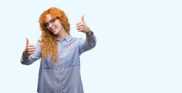 承認赤毛の若い公務員女性は笑顔と幸せな成功のため親指の手で肯定的なジェスチャーをしています。カメラ、勝者のジェスチャーを見てください。. - 写真・画像