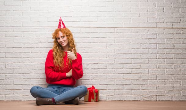 Mujer pelirroja joven sentada sobre la pared de ladrillo con sombrero de cumpleaños feliz con una gran sonrisa haciendo signo de ok, pulgar hacia arriba con los dedos, señal excelente
 - Foto, imagen