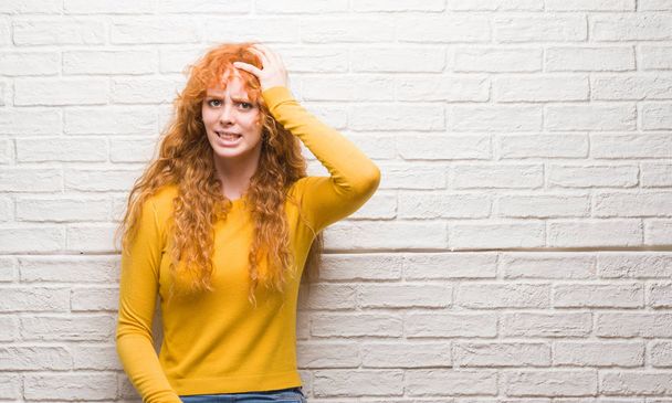 Young redhead woman standing over brick wall stressed with hand on head, shocked with shame and surprise face, angry and frustrated. Fear and upset for mistake. - Photo, Image