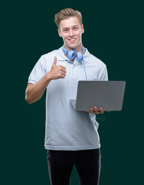 Genç yakışıklı sarışın adam ok işareti, başparmak ile parmak, mükemmel işareti yapıyor kocaman bir gülümseme ile mutlu bir dizüstü bilgisayar kullanarak - Fotoğraf, Görsel