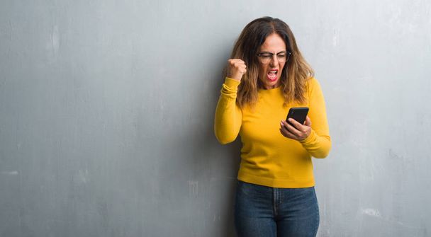 Mulher hispânica de meia idade usando smartphone irritado e frustrado gritando com raiva, louco e gritando com a mão levantada, conceito de raiva
 - Foto, Imagem