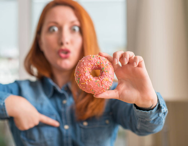 Pelirroja sosteniendo donut en casa con la cara sorpresa señalando el dedo a sí mismo
 - Foto, Imagen
