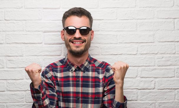 Young adult man wearing sunglasses standing over white brick wall screaming proud and celebrating victory and success very excited, cheering emotion - Photo, Image
