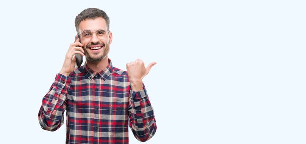 Young hipster adult man talking on the phone pointing and showing with thumb up to the side with happy face smiling - Foto, immagini