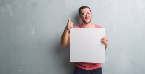 Homem caucasiano jovem sobre parede grunge cinza segurando um banner em branco feliz com grande sorriso fazendo sinal ok, polegar com os dedos, excelente sinal
 - Foto, Imagem
