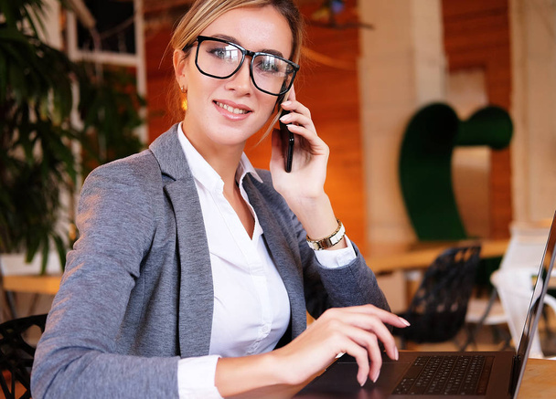 Good business talk. young beautiful woman in glasses talking on mobile phone and using laptop with smile while sitting at her working place. - Foto, afbeelding