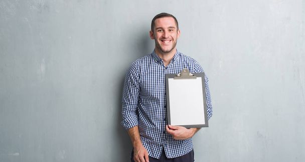 Молодий білий чоловік над сірою гранжевою стіною тримає буфер обміну з щасливим обличчям, що стоїть і посміхається з впевненою посмішкою, показуючи зуби
 - Фото, зображення