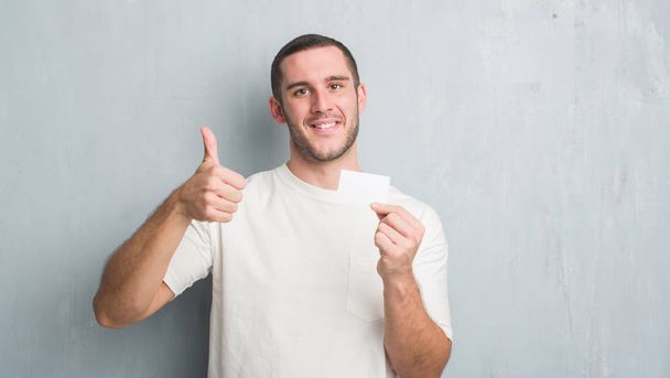 Homem caucasiano jovem sobre parede grunge cinza mostrando cartão de visita em branco feliz com grande sorriso fazendo sinal ok, polegar para cima com os dedos, excelente sinal
 - Foto, Imagem