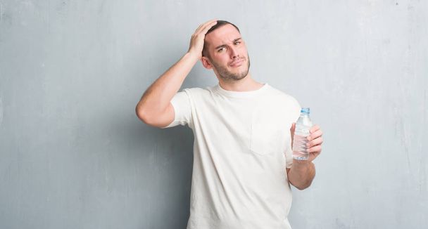 Young caucasian man over grey grunge wall holding bottle of water stressed with hand on head, shocked with shame and surprise face, angry and frustrated. Fear and upset for mistake. - Photo, Image