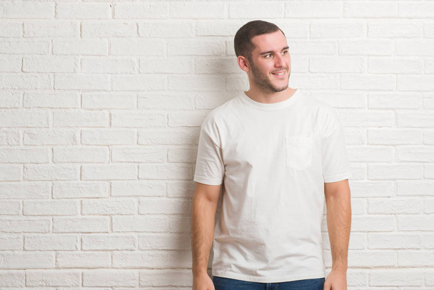 Young caucasian man standing over white brick wall looking away to side with smile on face, natural expression. Laughing confident. - Photo, Image