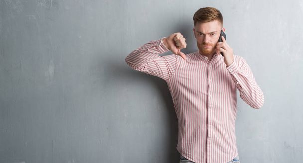 Genç Kızıl saçlı adam kızgın yüzü, eksi işareti aşağı antipati başparmak ile gösterilen, ret kavramı ile telefonda konuşurken gri grunge duvarın üzerinden - Fotoğraf, Görsel