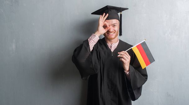 Νέοι κοκκινομάλλα άνδρα πάνω σε τοίχο γκρι grunge απόφοιτος στολή κρατώντας σημαία Γερμανίας με χαρούμενο πρόσωπο χαμογελά κάνει εντάξει σημάδι με το χέρι στο μάτι που αναζητούν μέσα από τα δάχτυλα - Φωτογραφία, εικόνα