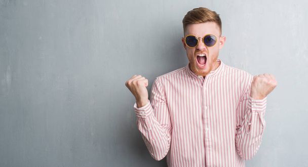Young redhead man over grey grunge wall wearing retro sunglasses annoyed and frustrated shouting with anger, crazy and yelling with raised hand, anger concept - Fotó, kép