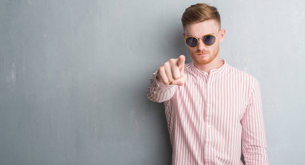 Молодой рыжий мужчина на серой гранж-стене в ретро-солнцезащитных очках, указывающих пальцем на камеру и на тебя, знак руки, позитивный и уверенный жест спереди
 - Фото, изображение