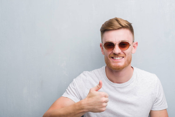 Молодой рыжий мужчина над серой гранж-стеной в ретро-солнцезащитных очках счастливый с большой улыбкой делает хорошо знак, палец вверх пальцами, отличный знак
 - Фото, изображение