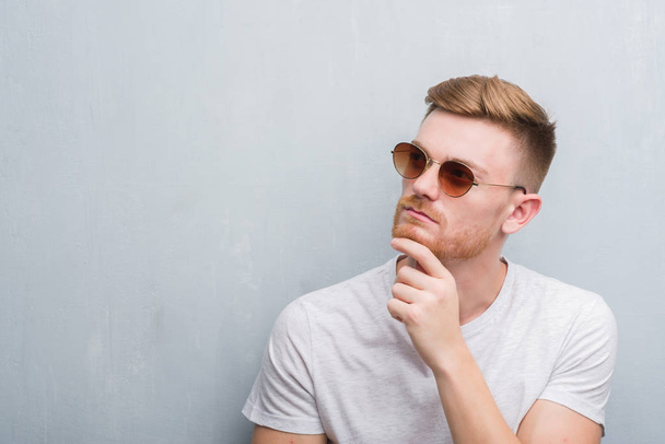 Jovem ruiva homem sobre cinza grunge parede vestindo óculos de sol retro rosto sério pensando em questão, ideia muito confusa
 - Foto, Imagem