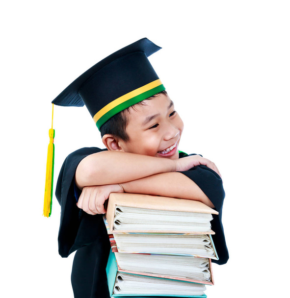 Knappe Aziatische kind in afstuderen jurk en pet met vele boeken. Gelukkig jongetje glimlachend met perfecte glimlach op de dag van zijn afstuderen. Geïsoleerd op witte achtergrond. - Foto, afbeelding