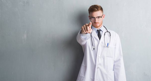 Nuori punapää lääkäri mies harmaan grunge seinä osoittaa sormella kameraan ja sinulle, käsi merkki, positiivinen ja luottavainen ele edestä
 - Valokuva, kuva