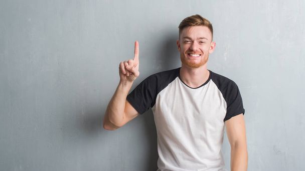 Mladá zrzka muž přes zeď šedá grunge ukazující a směřující nahoru s prstem číslo jedna, zatímco s úsměvem a spokojené. - Fotografie, Obrázek