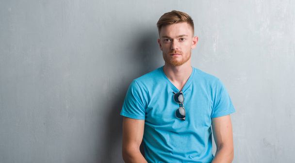Young redhead man over grey grunge wall wearing casual outfit with serious expression on face. Simple and natural looking at the camera. - Photo, Image