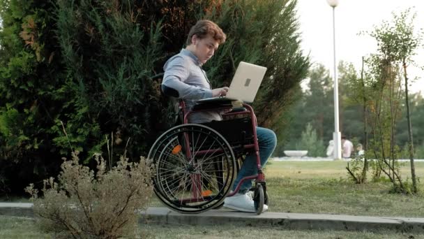 Un hombre discapacitado está sentado en una silla de ruedas y trabajando en un portátil en el parque
 - Metraje, vídeo