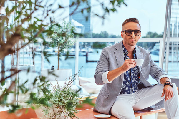 Pewny siebie człowieka sukcesu z stylowe włosy, ubrany w eleganckie nowoczesne ubrania posiada kawę siedząc na stole w kawiarni na świeżym powietrzu na tle nabrzeża. - Zdjęcie, obraz