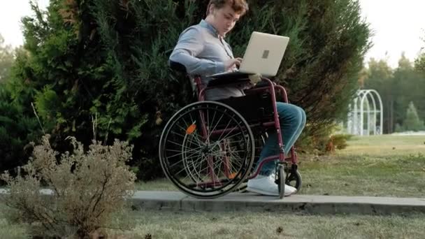 Niepełnosprawny mężczyzna siedzi na wózku inwalidzkim i działa na laptopie w parku - Materiał filmowy, wideo