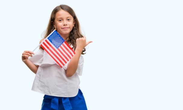 Bruna ragazza ispanica che tiene la bandiera degli Stati Uniti d'America puntando e mostrando con il pollice verso l'alto sul lato con il viso felice sorridente
 - Foto, immagini
