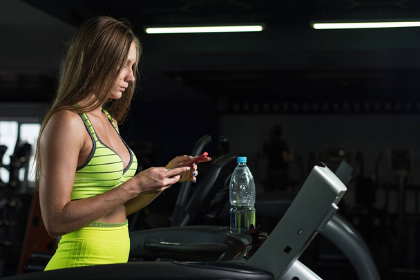 Beautiful sporty young Caucasian girl running on a treadmill holding her mobile phone in the gym - Φωτογραφία, εικόνα