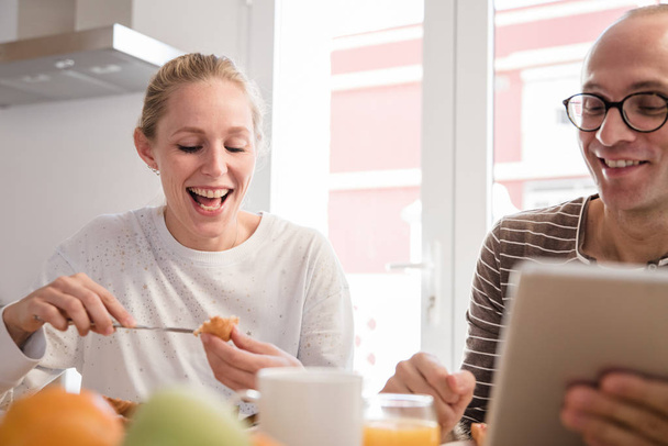 Couple laughing as they eat breakfast of fruit and croissants while sit at a table looking at a pc tablet - Foto, Imagem