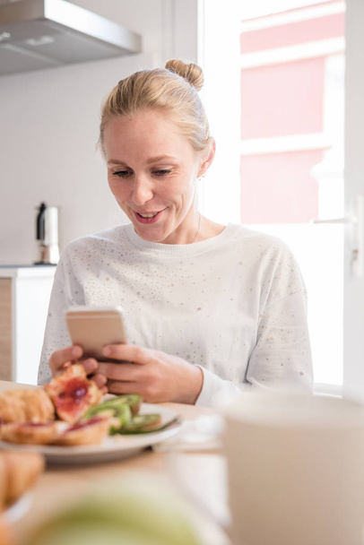 Woman sitting at a table and looking down at her phone as she smiles at breakfast - Foto, immagini