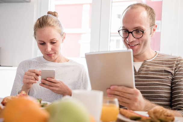 Couple looking down at their devices while sitting at a table and eating a breakfast - Foto, Imagem