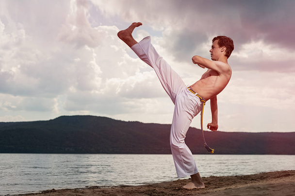 Young guy trains capoeira on sky backround. Concept about people, lifestyle and sport. A men performs martial the kick - Φωτογραφία, εικόνα