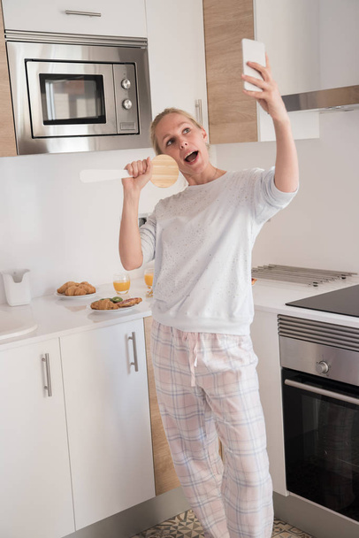 Женщина делает селфи на кухне, когда держит кухонную утварь и делает вид, что это микрофон
 - Фото, изображение