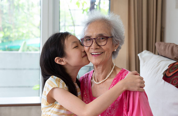 Портрет милой маленькой девочки и ее прекрасной бабушки, сидящей дома на диване, азиатской девушки, целующей свою улыбающуюся бабушку в щеку
 - Фото, изображение
