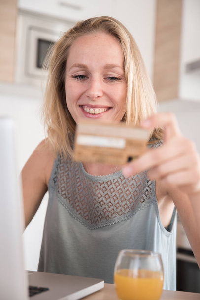 Woman sitting at a table in a kitchen as she holds her credit card in her hand and smiles - Fotoğraf, Görsel