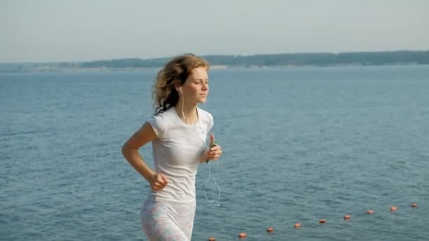 A young woman with a fine figure is engaged in gymnastics in the sea at dawn. She runs along the seacoast in headphones, Super slow motion - Video