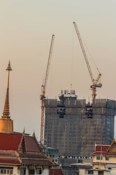 Τα Πύργος γερανός φλόκων σε συγκυριαρχία εργοτάξιο πάνω από χάλυβα πλαίσιο ανάμεσα σε πολυόροφο κτήριο. Γερανοί ουρανοξύστης κτίσμα με τον πύργο στην κορυφή κάτω από το δραματικό ουρανό φόντο. - Φωτογραφία, εικόνα