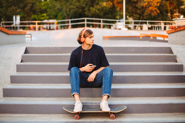 Joven patinador en auriculares naranjas con teléfono móvil en las manos sentado en las escaleras en el moderno parque de skate
 - Foto, Imagen